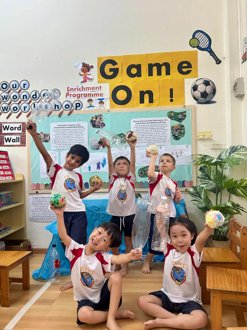 Five Shaws preschoolers holding up a recycling craft project in front of their classroom display board