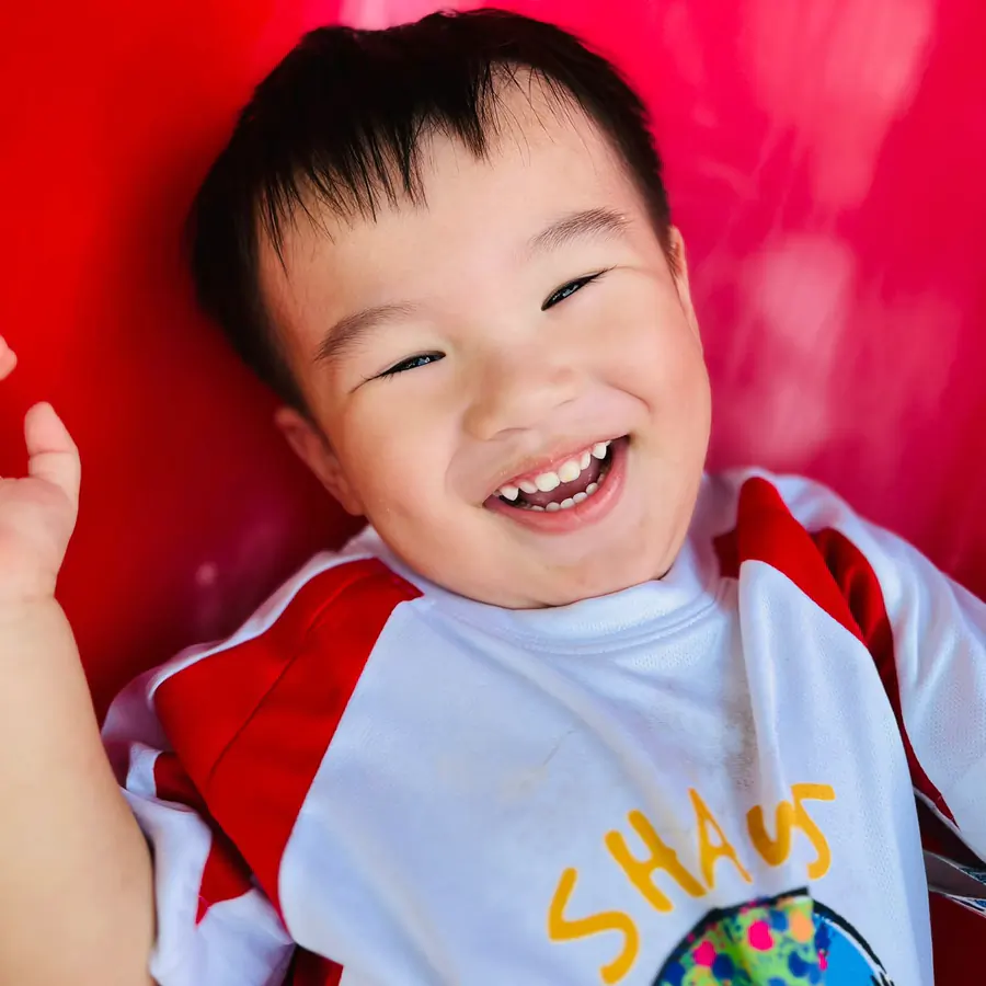 A boy in a Shaws Preschool uniform beaming at the camera during outdoor play