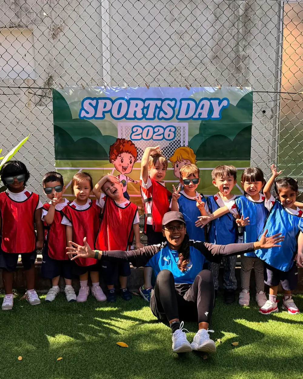 A Shaws Preschool class in red and blue team bibs cheering with their coach under a Sports Day banner