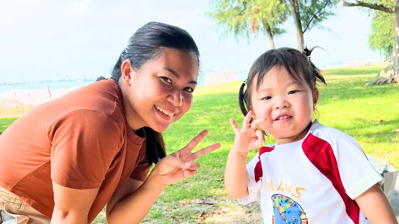 A Shaws Preschool teacher and a toddler crouching on the grass by the sea, both making peace signs at the camera